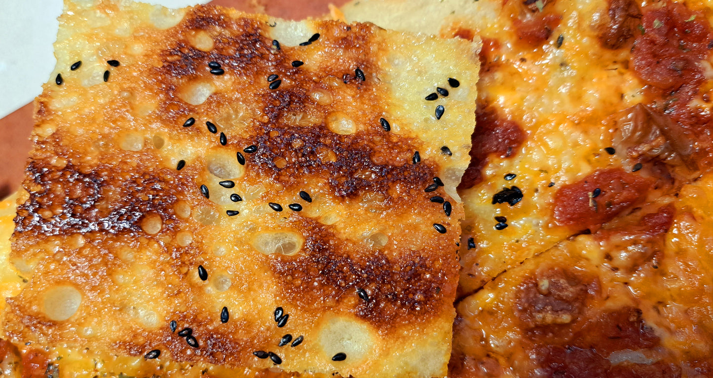 The bottom exterior of the Grandma's Pizza; Showing a golden exterior with black toasted sesame seeds for extra crunch. Compliments well with the sweet and sour pomodoro sauce and melty cheese.