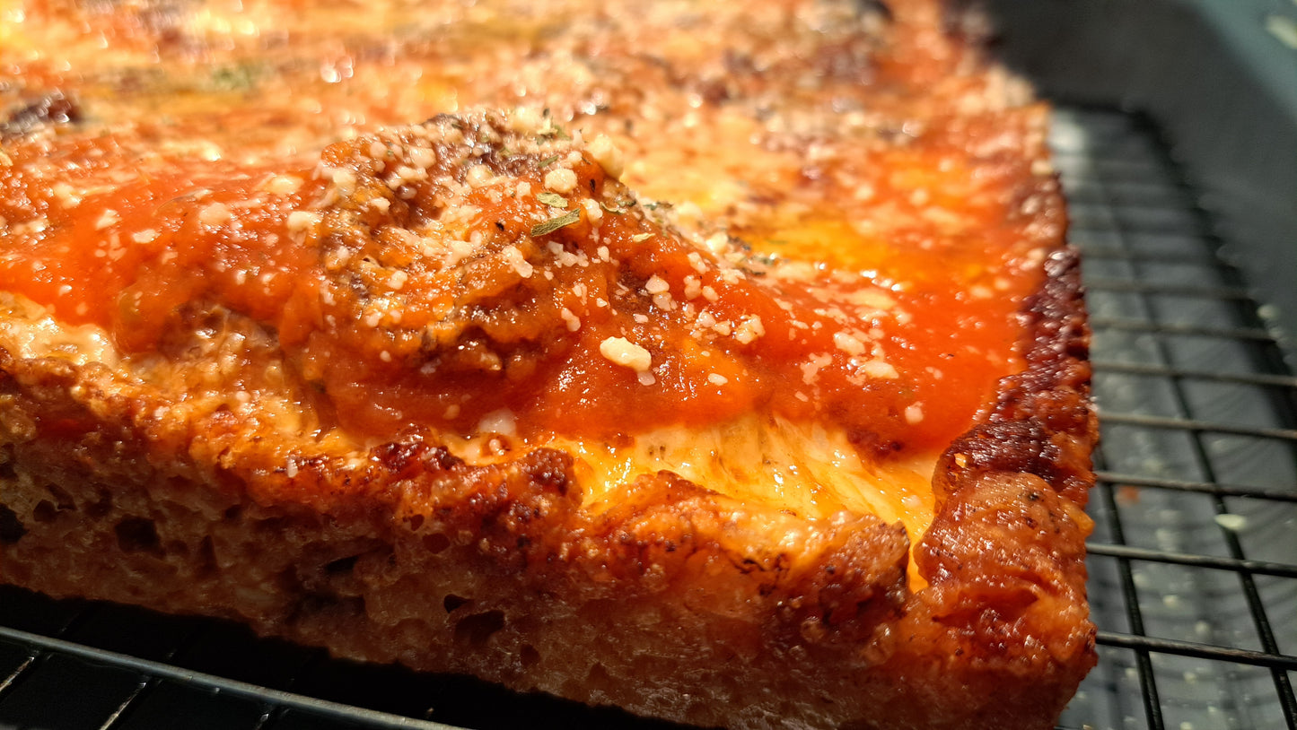 A close-up of a freshly baked Detroit-style pizza, showcasing its distinctive characteristics. The pizza has a thick, rectangular crust that is golden brown and crispy on the edges. The top is generously covered with a rich tomato sauce, which glistens and indicates a flavorful base. Additionally, there is a sprinkle of grated cheese and possibly herbs on top, adding to the visual appeal and flavor profile. The crust appears airy and well-cooked, with a nice texture that suggests it's both crunchy and soft