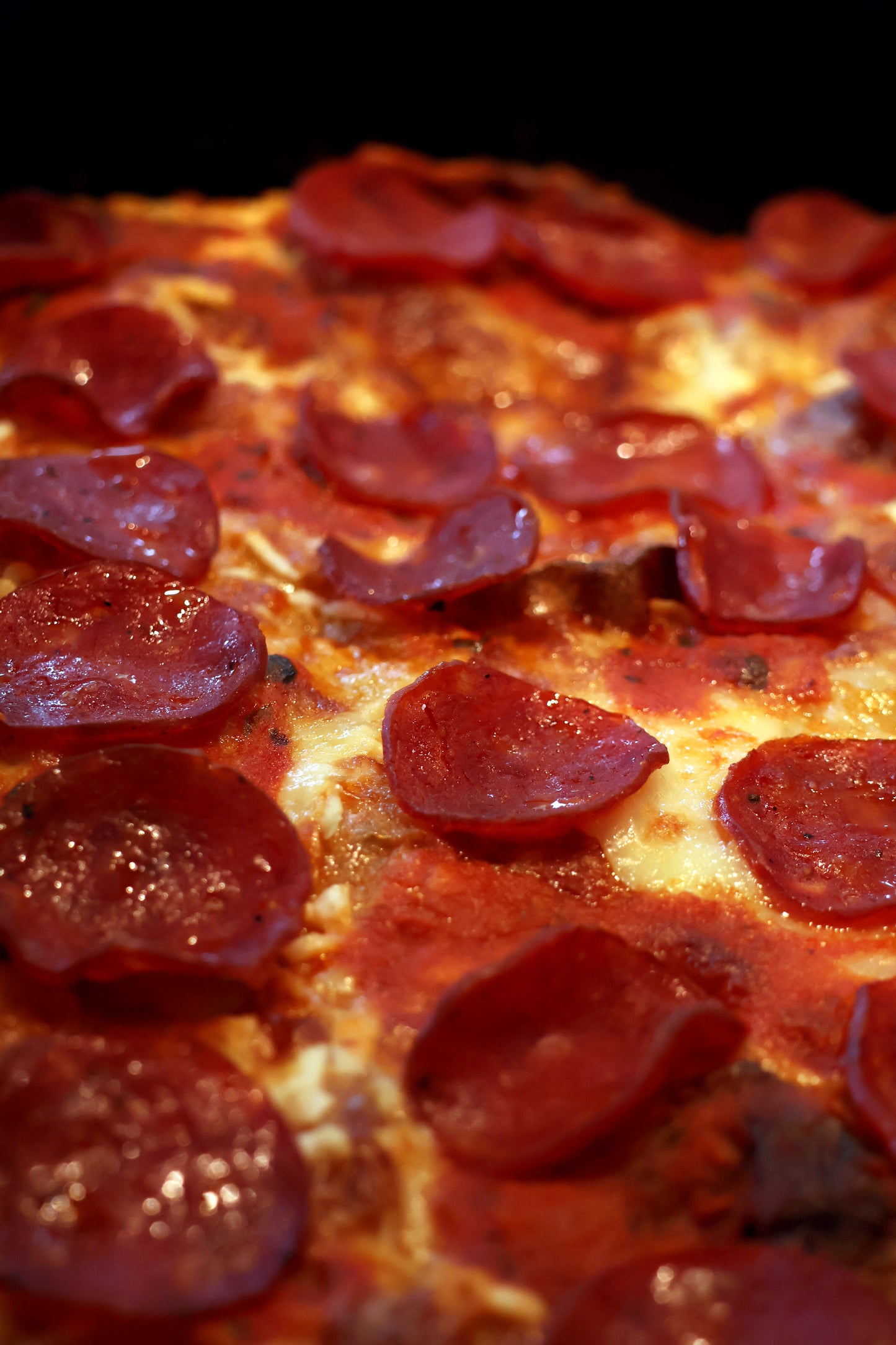 A delicious-looking pepperoni Detroit-style pizza. The pizza features a thick, rectangular crust that is crispy on the outside and soft inside. It is topped generously with slices of pepperoni, which have a slightly crisped edge, indicating they were baked to perfection. The cheese appears to be melted and gooey, likely extending all the way to the edges, creating a caramelized cheese crust. The sauce is not prominently visible but is likely layered beneath the cheese and pepperoni, characteristic of Detroi