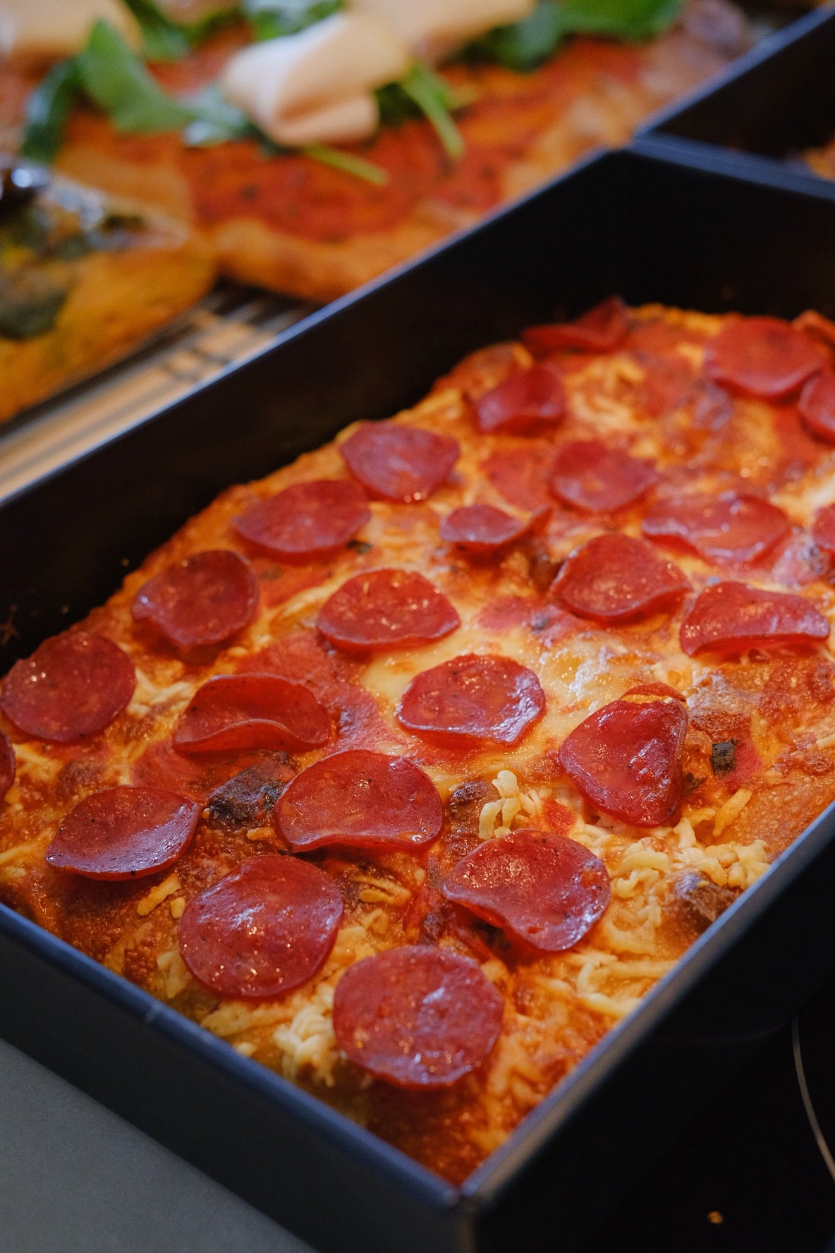 A rectangular pan of freshly baked pizza, prominently featuring slices of pepperoni on top. The pizza has a golden-brown crust and a generous layer of melted cheese, which appears to be bubbling slightly, indicating it's hot and fresh out of the oven.