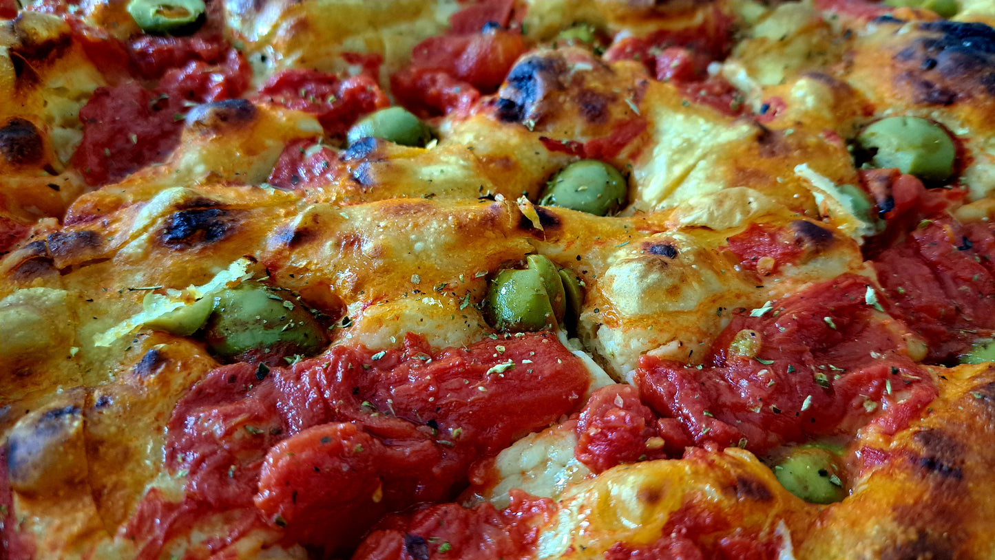 Focaccia that is topped with a rich tomato sauce and green olives. The crust is golden and crispy, with some charred spots, indicating it has been baked at a high temperature. The vibrant red of the tomato sauce contrasts with the green olives, which are scattered throughout the topping. The dish is seasoned with herbs, possibly oregano or basil, adding depth to the flavor profile.