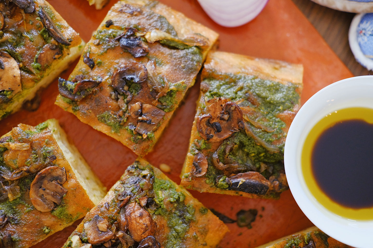 focaccia-style bread topped with sautéed mushrooms and a vibrant green sauce, possibly pesto. The bread is cut into rectangular pieces, revealing a fluffy, golden crust with a crispy surface. In addition to the mushrooms, the green sauce adds a fresh and herbaceous flavor, enhancing the overall taste.