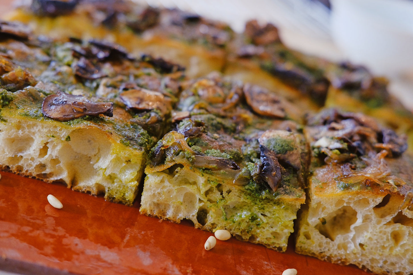 Focaccia topped with sautéed mushrooms and a vibrant green sauce, likely pesto. The focaccia has a golden-brown crust and a soft, airy interior, with visible holes that indicate a well-aerated dough.