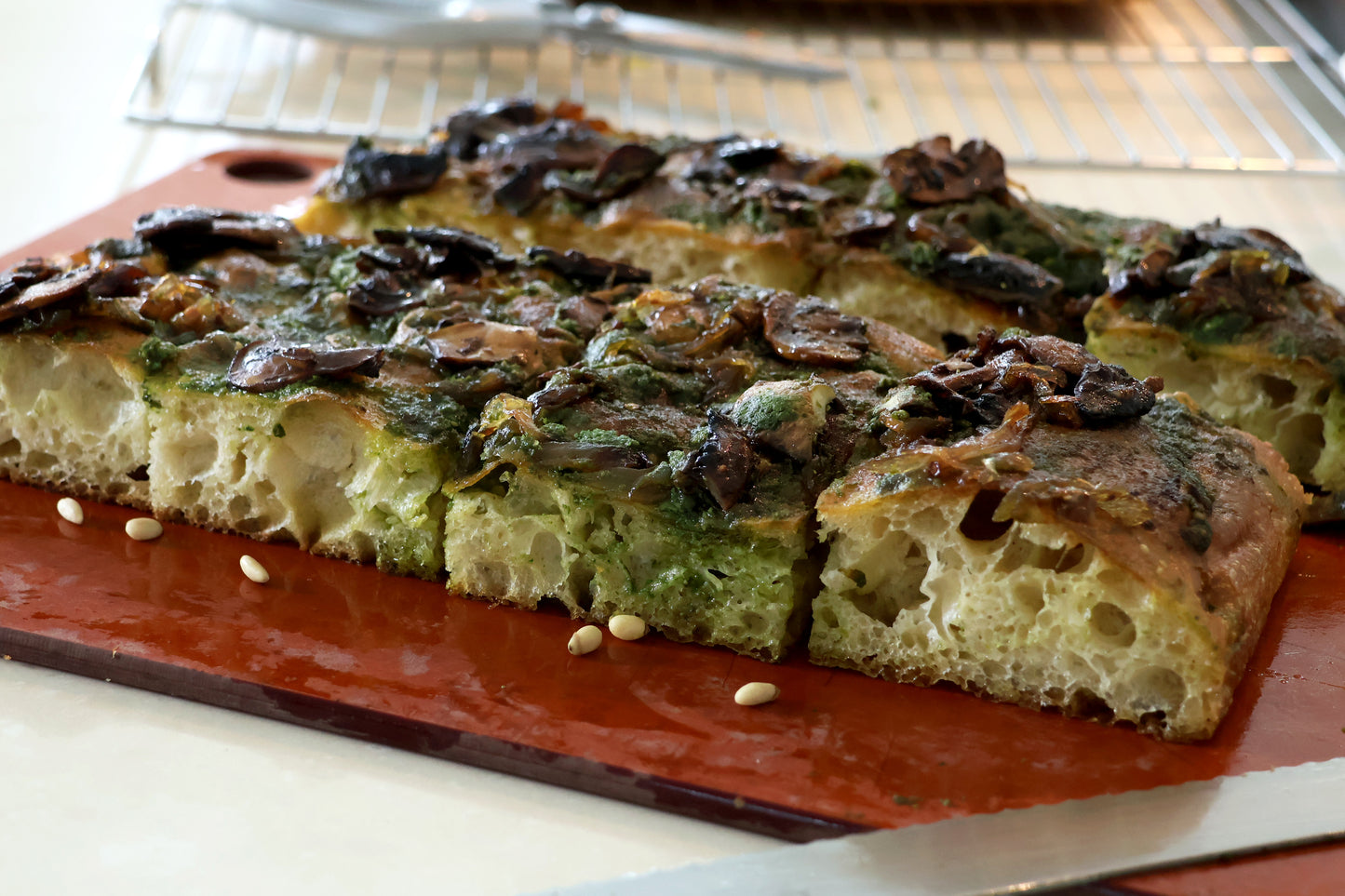 A close-up view of a slice of focaccia, showcasing its soft, airy interior and golden-brown crust. The focaccia is topped with sautéed mushrooms and a vibrant green sauce, likely pesto, which adds both flavor and visual appeal. With large holes indicating a well-aerated dough, contributing to its lightness. Pine nuts are scattered on the surface, adding a touch of elegance and additional flavor.