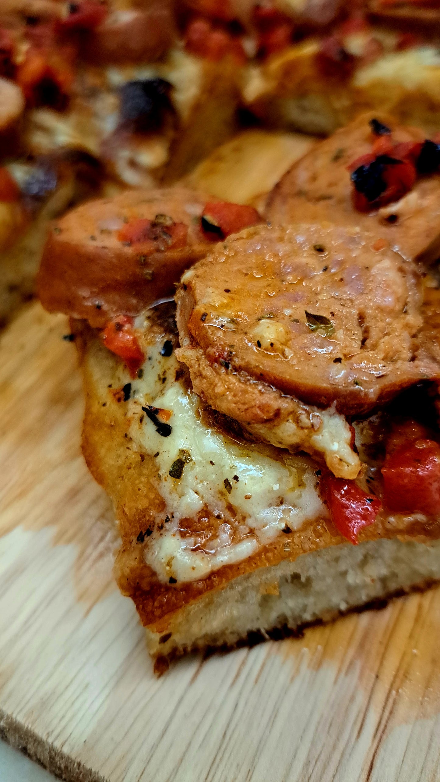 A close-up view of a rectangular Detroit pizza topped with slices of sausage and a rich, chunky sauce. The crust appears thick and golden, indicating a crispy texture.
