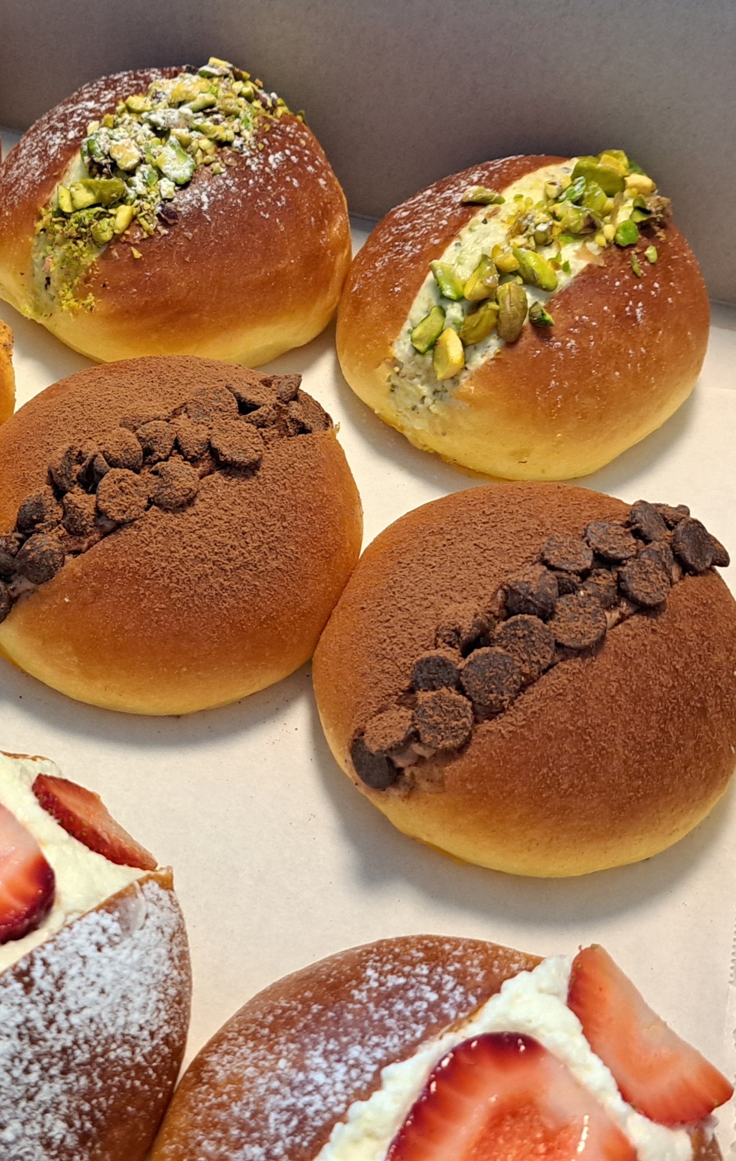 An assortment of maritozzi, traditional Italian sweet buns. Strawberry Topped: Some buns are filled with whipped cream and topped with fresh strawberry slices, adding a fruity freshness.
Pistachio: One bun features a green pistachio filling or topping, providing a nutty flavor.
Chocolate Variants: Several buns are adorned with chocolate chips or cocoa powder, appealing to chocolate lovers.