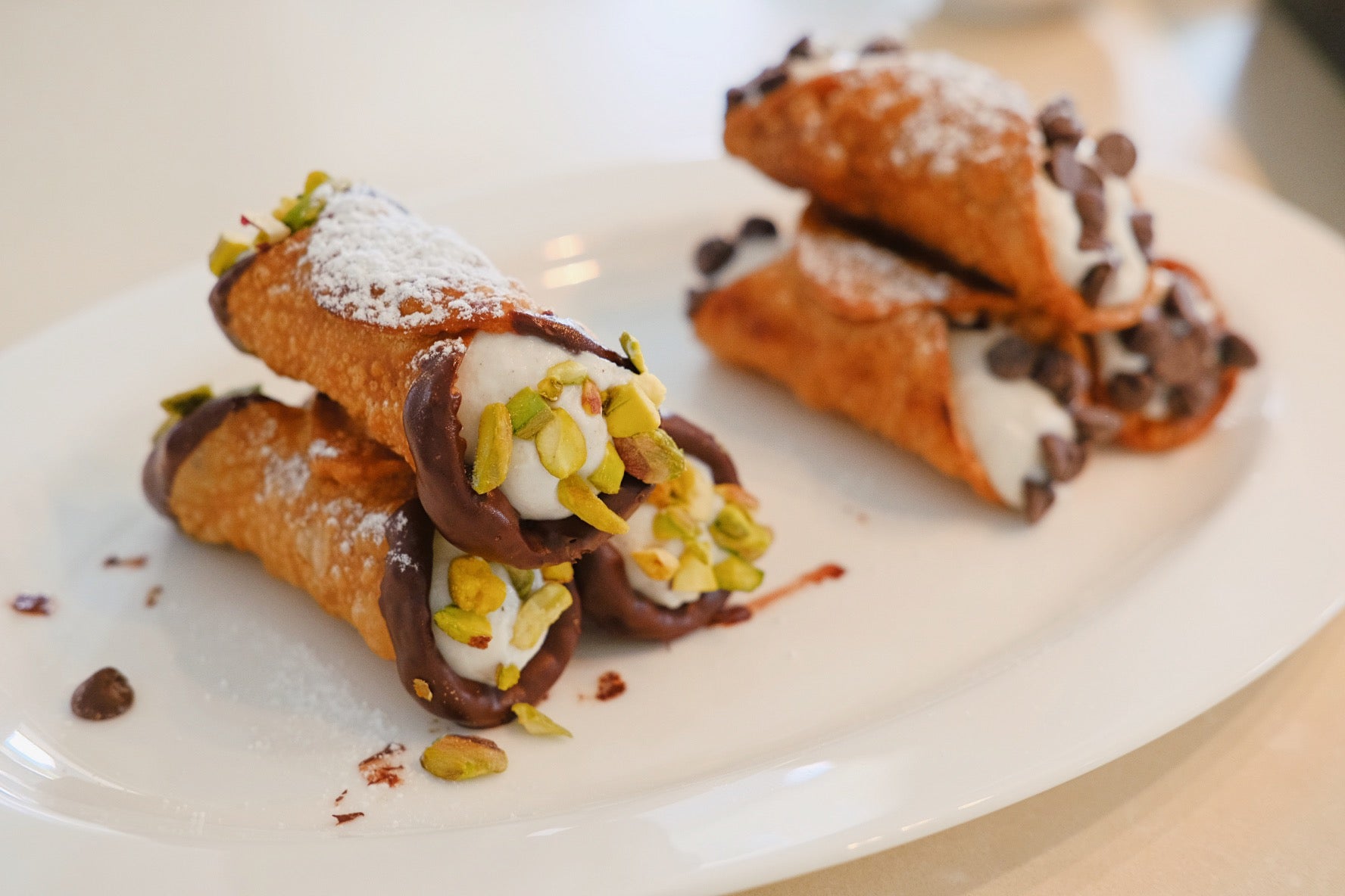 On the left, deep fried cannoli (Pasta shells), filled with ricotta cream with the ends coated in dark chocolate and grounded and toasted pistachios. On the right, deep fried pasta shells filled with ricotta cream and topped with chocolate chips on the ends. Dusted with icing sugar.