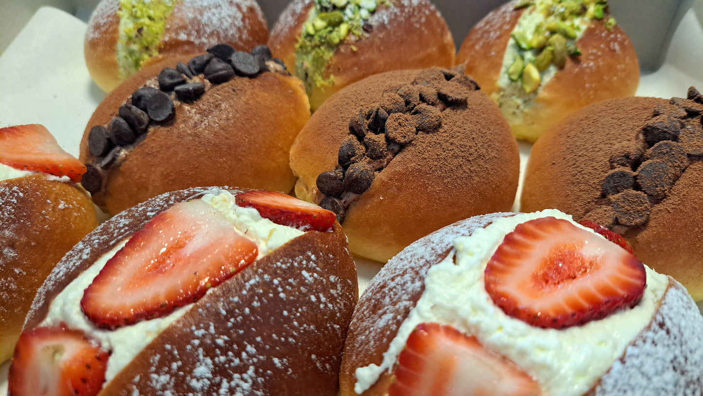 An assortment of maritozzi, traditional Italian sweet buns. Strawberry Topped: Some buns are filled with whipped cream and topped with fresh strawberry slices, adding a fruity freshness.
Pistachio: One bun features a green pistachio filling or topping, providing a nutty flavor.
Chocolate Variants: Several buns are adorned with chocolate chips or cocoa powder, appealing to chocolate lovers.