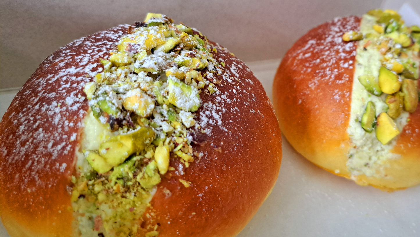 Two delicious-looking maritozzi, which are traditional Italian sweet buns. Pistachio flavour; with the contrast of the green pistachios against the golden bread and white powdered sugar, making them look appetizing and ready to enjoy.